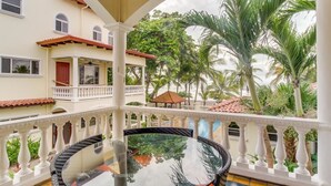 Uma varanda com mesa de vidro e cadeiras de vime, com vista para um jardim com palmeiras e um edifício com telhado vermelho.