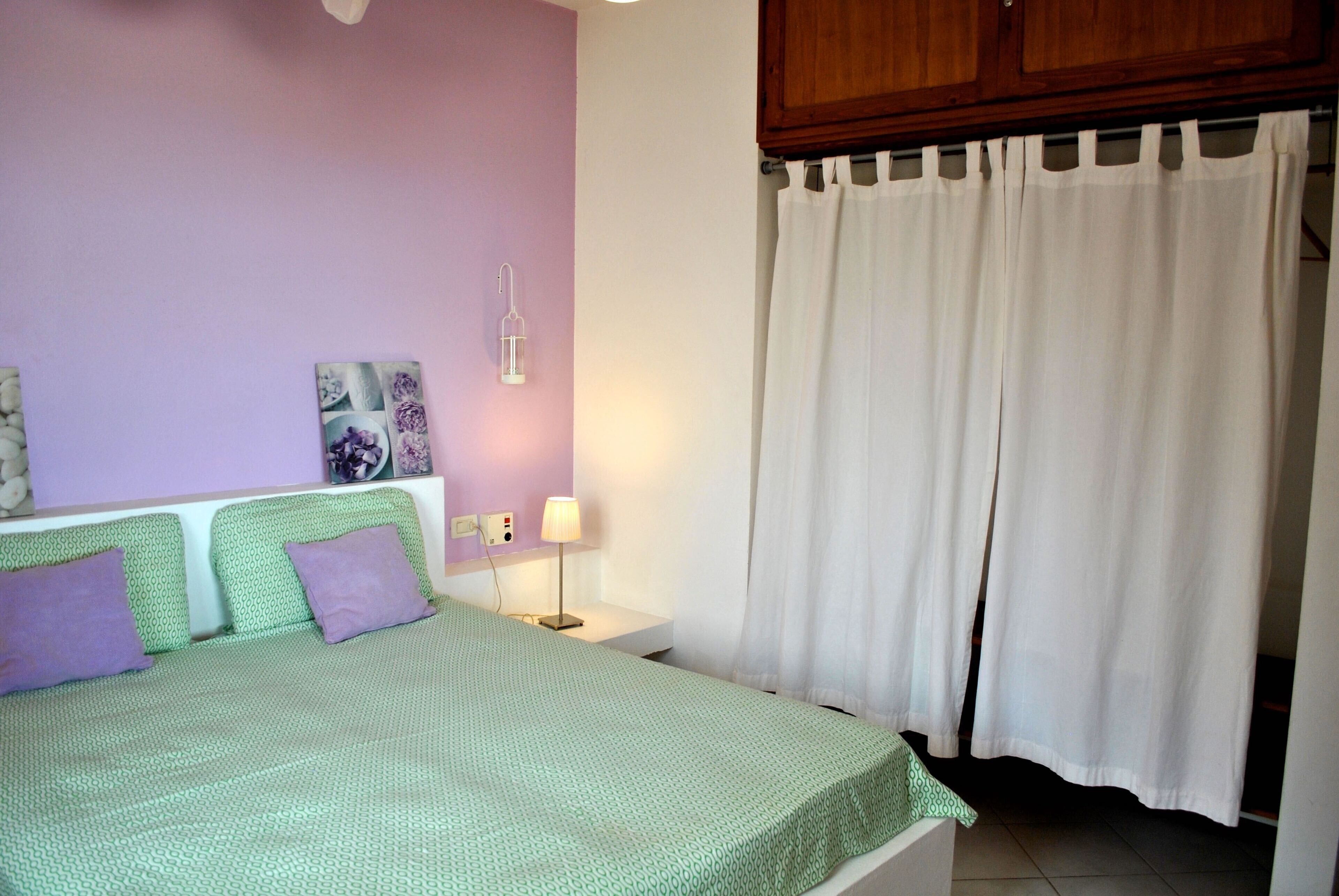 Un dormitorio con cubrecama verde, almohadas moradas, una mesita de noche blanca con lámpara, una luz empotrada en la pared y una cortina blanca.