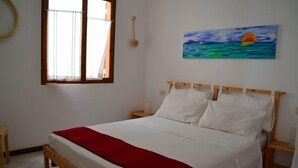 A bedroom with a wooden bed, white bedding, a red throw blanket, a painting of a sunset over the ocean, and a window with curtains. - Sea View Apartments Boa Vista (Boa Vista)