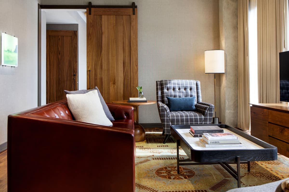 A modern hotel room with a leather sofa, a plaid armchair, a small side table with a lamp, and a television.