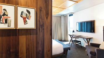 A hotel room with a bed, a desk, a chair, a TV, and two framed artworks on the wall.