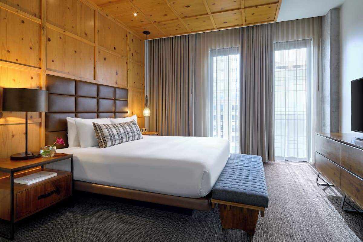 A hotel room with a large bed, a wooden headboard, a bedside table with a lamp, a bench, and a TV on a dresser.