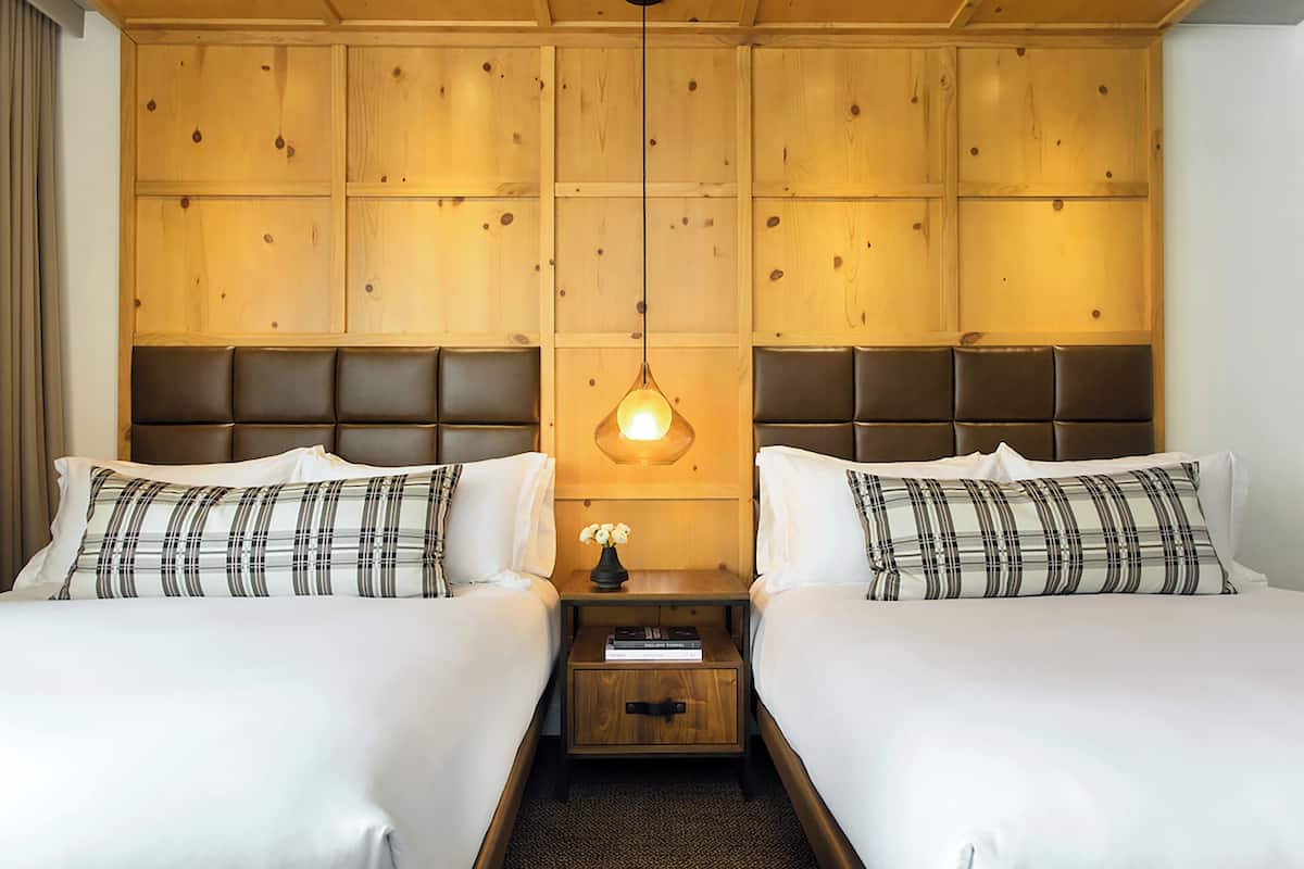 A hotel room with two beds, a wooden headboard, a nightstand with a lamp, and a small table with a book.