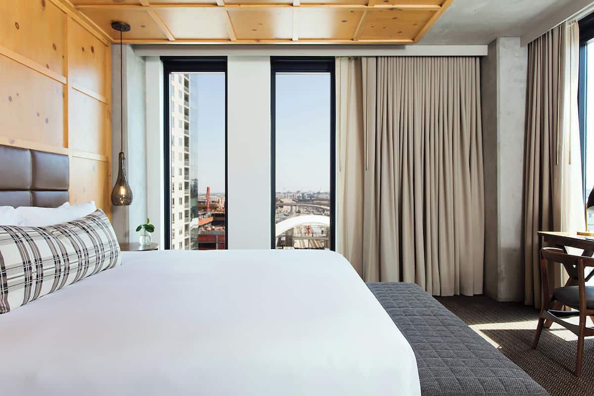 A hotel room with a large bed, a wooden headboard, a desk with a chair, and a view of the city through large windows.