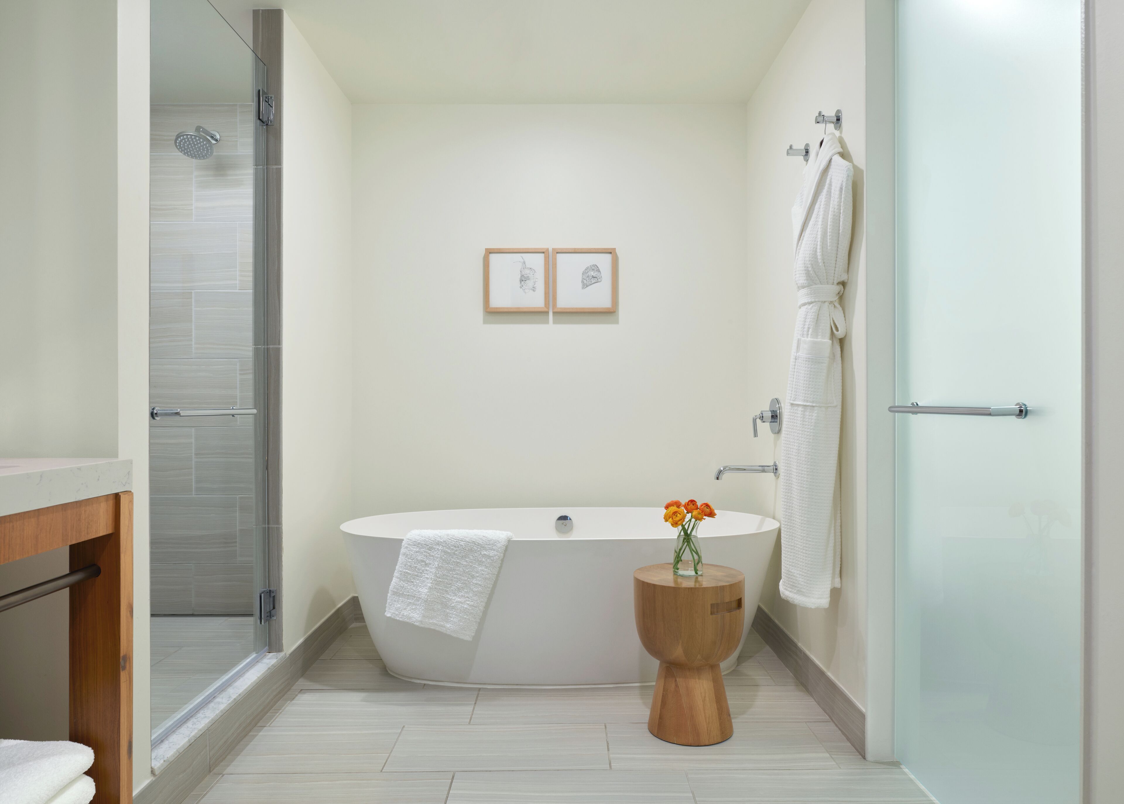 Ein modernes Badezimmer mit freistehender Badewanne, einer Duschkabine aus Glas und einem Holztischchen mit Blumen.'