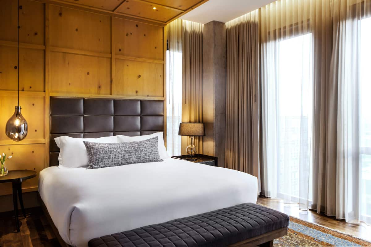 A hotel room with a large bed, a headboard, a bedside table with a lamp, and a view of the city through large windows.