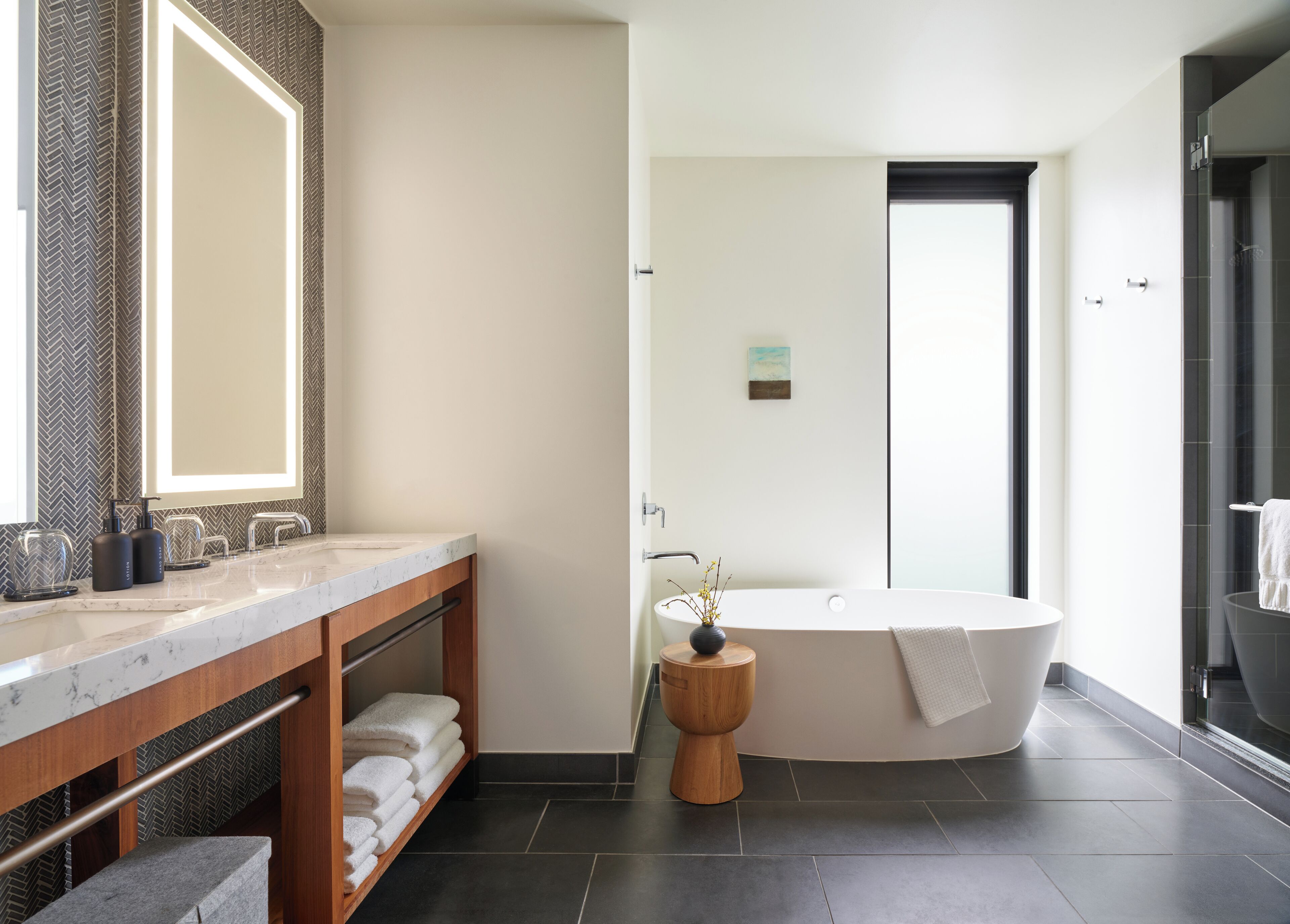 A modern bathroom with a large bathtub, a vanity with a mirror, and a wooden shelf with towels.