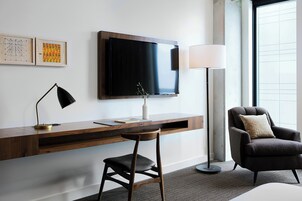 A modern hotel room with a wooden desk, a flat-screen TV, a floor lamp, and a comfortable armchair.
