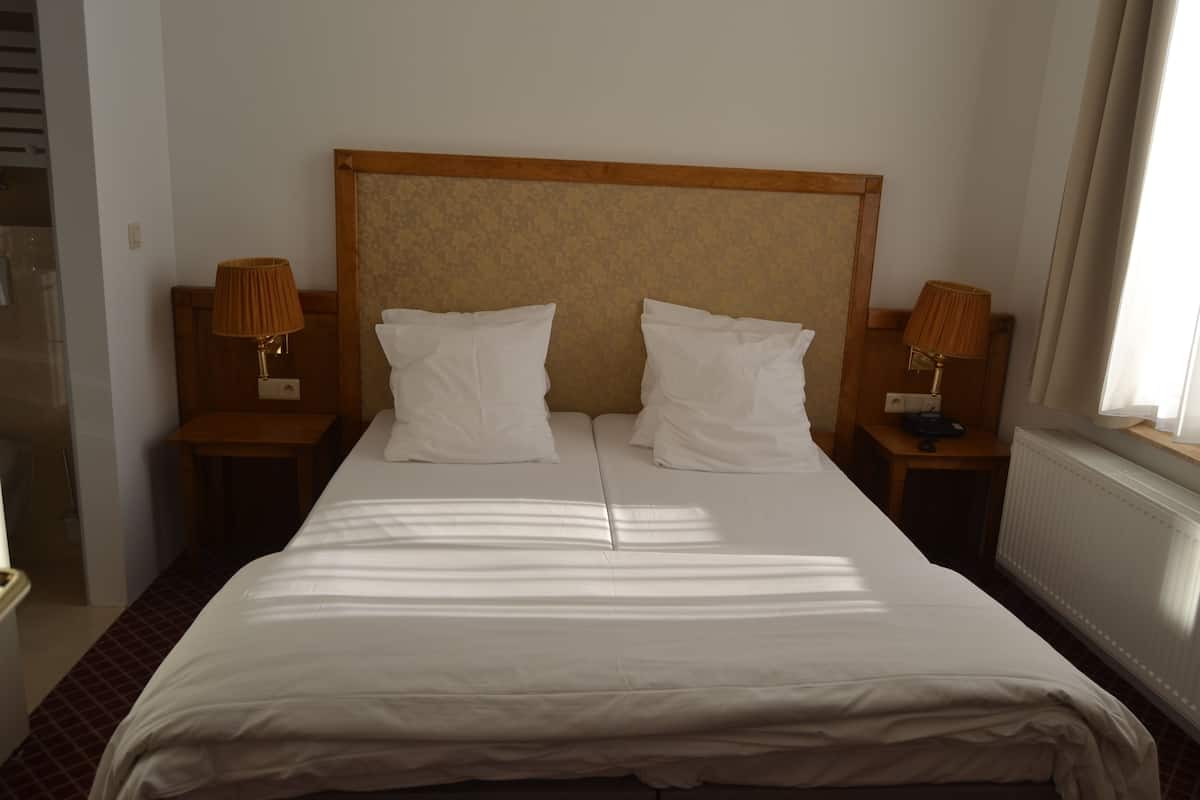 Een hotelkamer met een groot bed, twee nachtkastjes met lampen, een houten hoofdeinde en een raam met gordijnen.