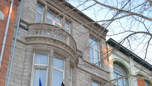 Ein historisches Gebäude mit einer steinernen Fassade, großen Fenstern und einem schwarzen schmiedeeisernen Zaun. Vor dem Gebäude werden Flaggen gezeigt.