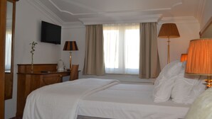 Ein Hotelzimmer mit einem großen Bett, einem Schreibtisch mit Lampe, einem an der Wand befestigten Fernseher und einem Fenster mit Vorhängen.