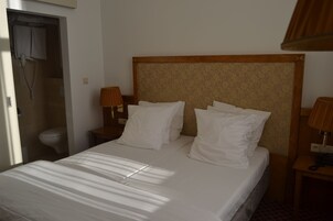 Una camera d'albergo con un letto, una testiera in legno, un comodino con una lampada e un bagno con il water e una doccia.