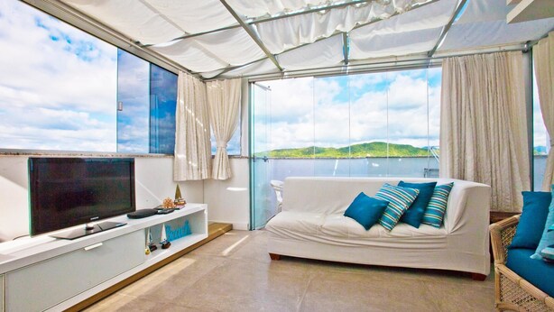 A modern living room with a large window, a flat-screen TV, a white sofa with blue cushions, and a wicker chair. - FL014 3 Bedroom Apartment By Senstay (Florianópolis)