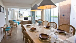 A dining area with a long table set for six, surrounded by wicker chairs, with a view of the outdoors through large windows. - FL014 3 Bedroom Apartment By Senstay (Florianópolis)