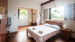 A bedroom with a wooden bed, a television on a stand, a window with curtains, and a small bedside table with a lamp. - FL023 6 Bedroom Holiday home By Senstay (Florianópolis)