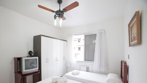 A hotel room with two single beds, a ceiling fan, a TV on a wooden stand, a window with curtains, and a framed picture on the wall.