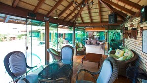 A covered outdoor seating area with wicker furniture and glass tables, overlooking a pool and garden. - FL004 5 Bedroom Villa By Senstay (Florianópolis)