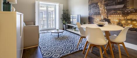A modern dining area with a round table, white chairs, a patterned rug, and a cityscape mural on the wall.