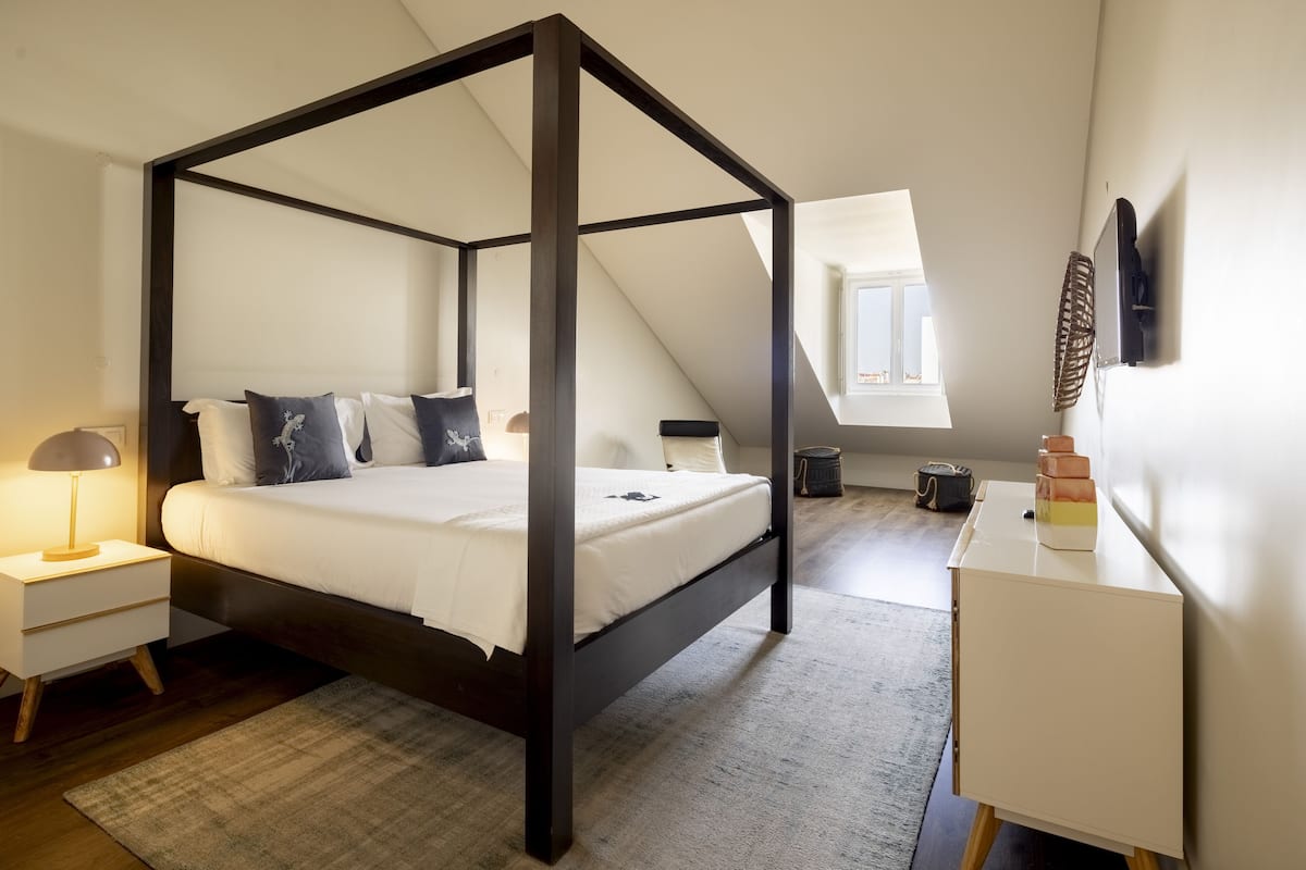 Ein Bett mit Baldachin, dessen Gestell dunkel ist, ein Nachttisch mit Lampenschirm in Weiß sowie ein kleiner Schreibtisch mit Ventilator.