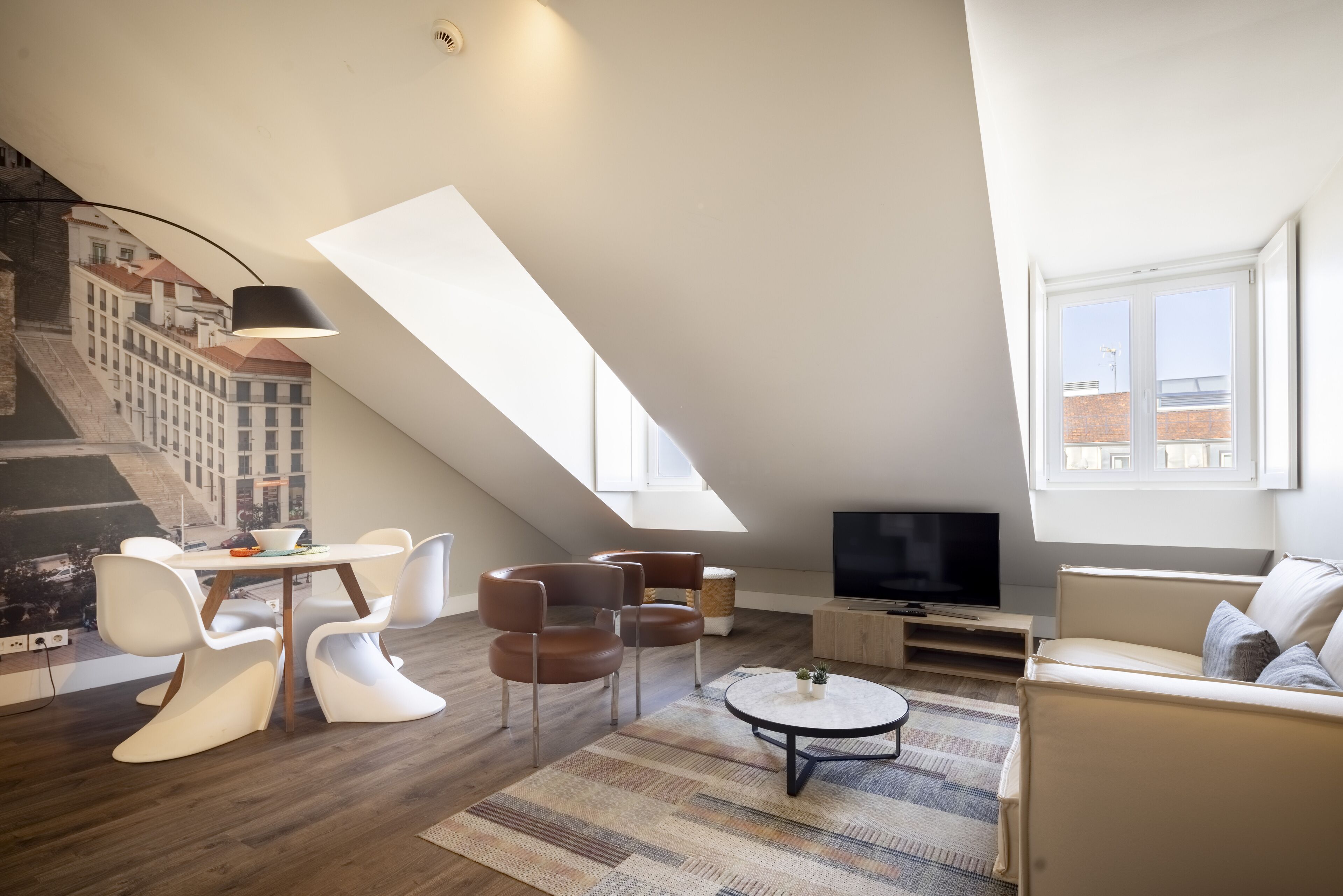 Un moderno loft con soffitto spiovente, zona pranzo con sedie bianche e marroni, tavolino rotondo, televisore a schermo piatto e un'ampia finestra con vista sugli edifici.