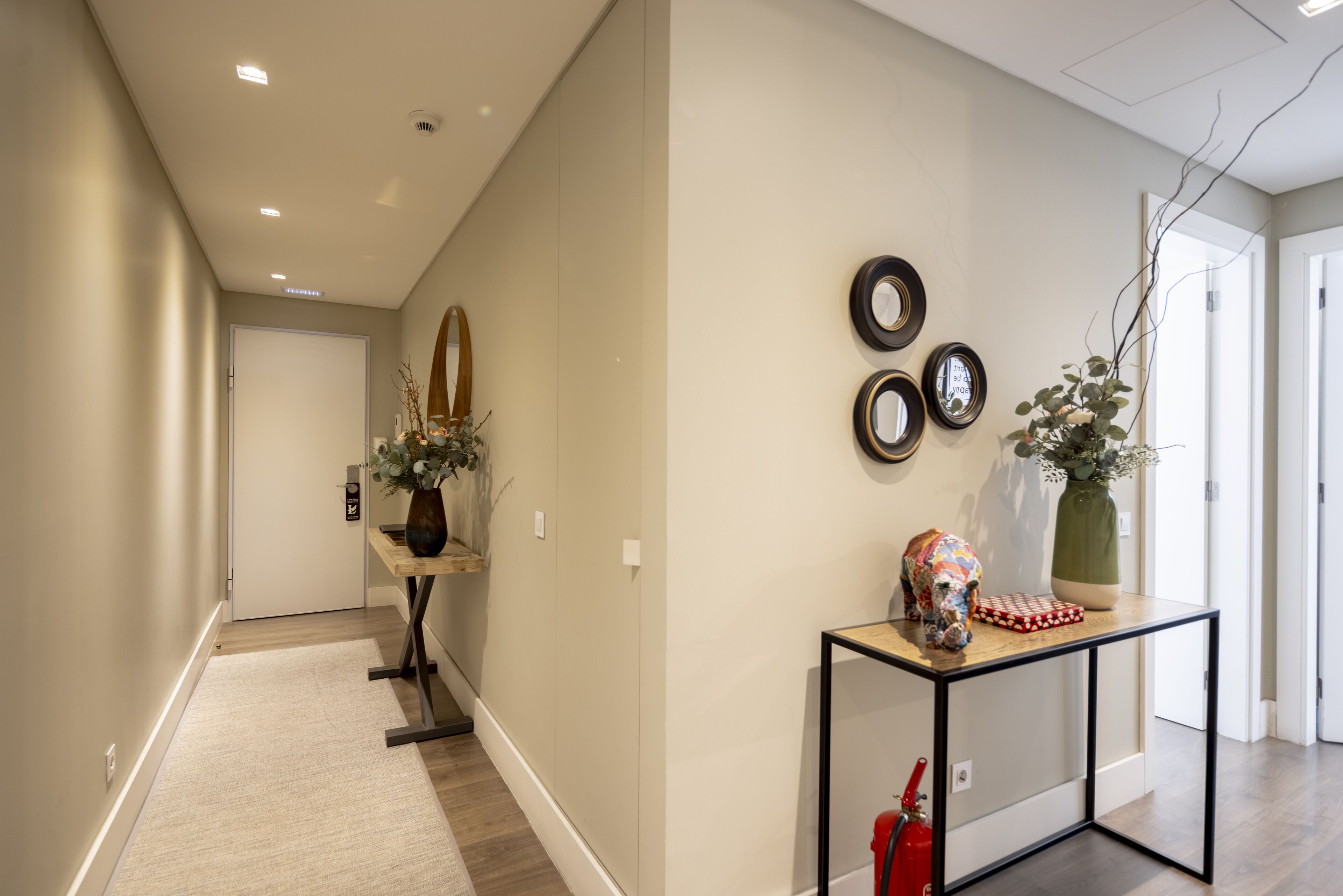 A hallway with a wooden floor, a small table with a vase and a decorative item, and three framed mirrors on the wall.