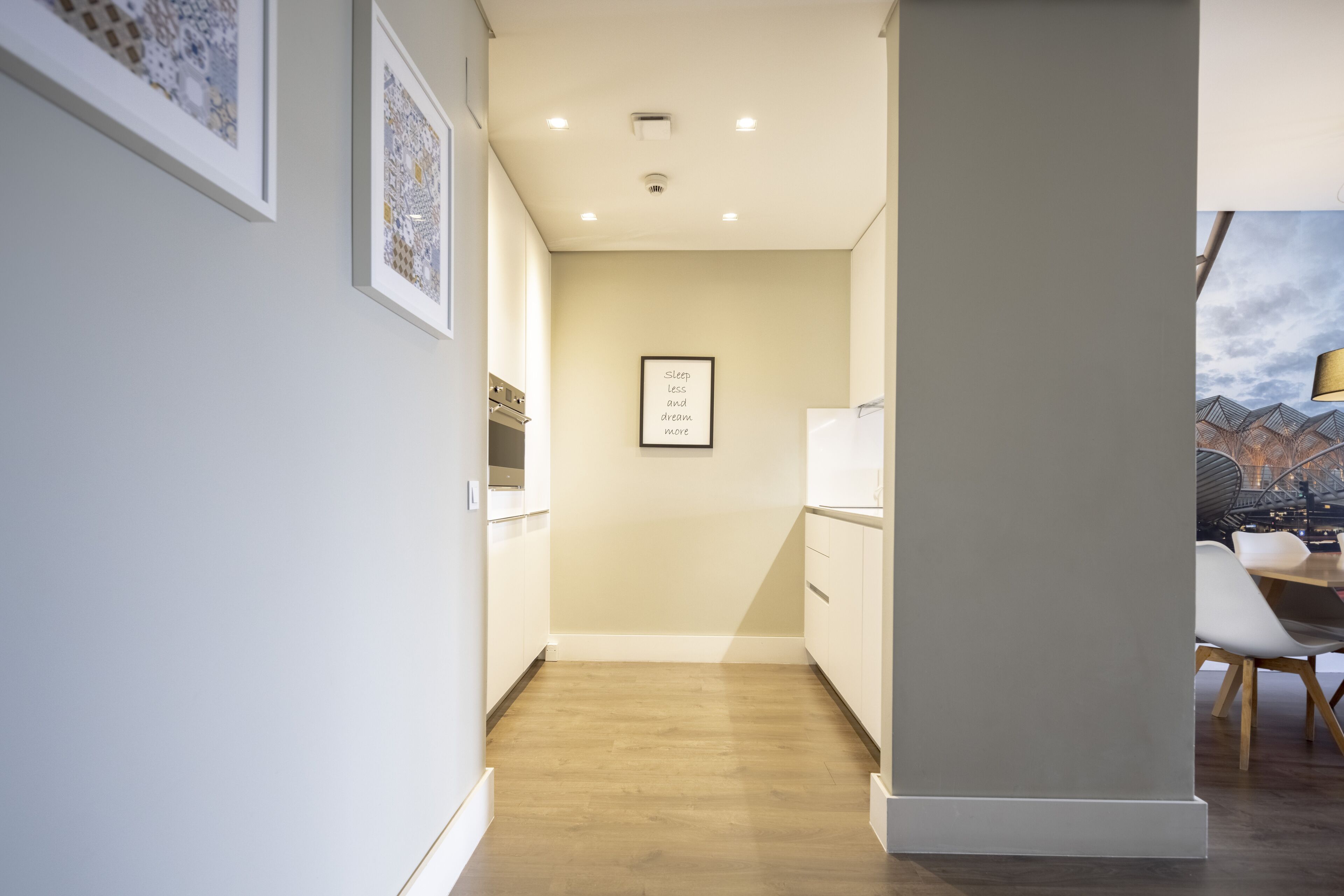 A narrow hallway with wooden flooring, white walls, and framed artwork. There is a kitchen area with cabinets and a refrigerator visible in the background.