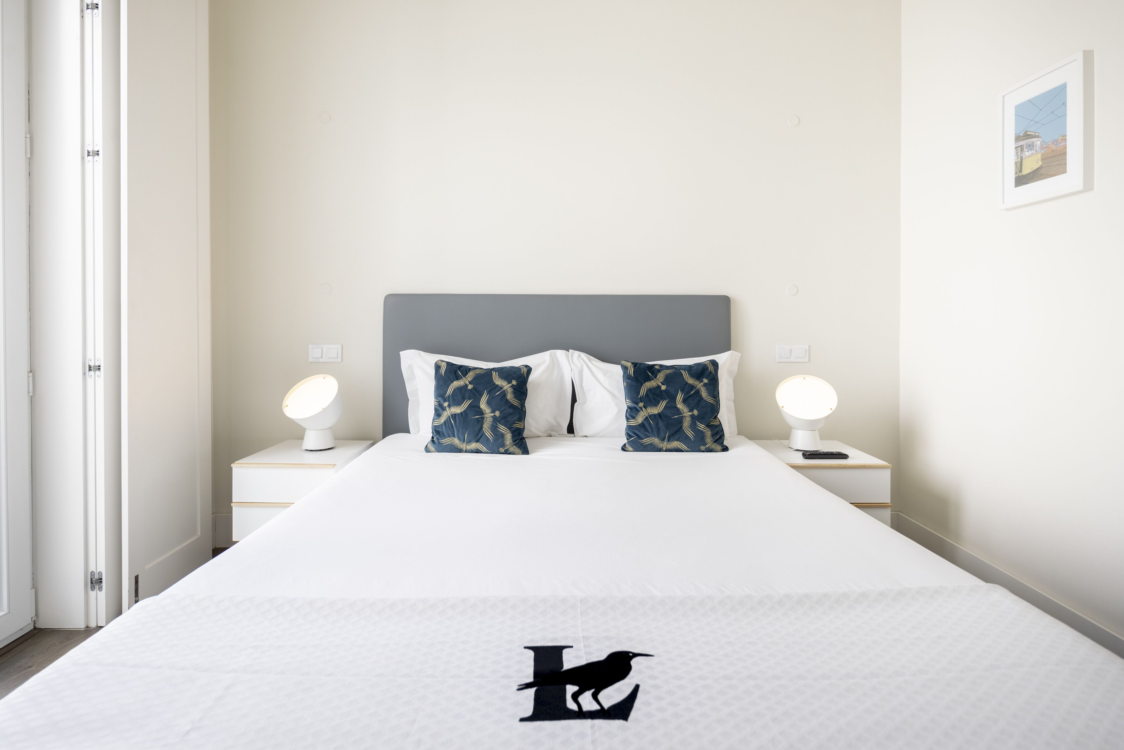 A neatly made bed with a white bedspread and a black bird logo, two matching pillows, two bedside lamps, and a framed picture on the wall.