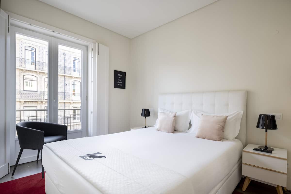 Ein Hotelzimmer mit einem großen Bett, zwei Nachttischen, einem Sessel und Blick auf Gebäude durch das Fenster.