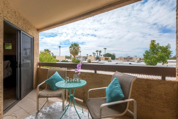 Un balcón con dos sillas de mimbre, una mesita redonda y vistas a una zona residencial con palmeras y casas.