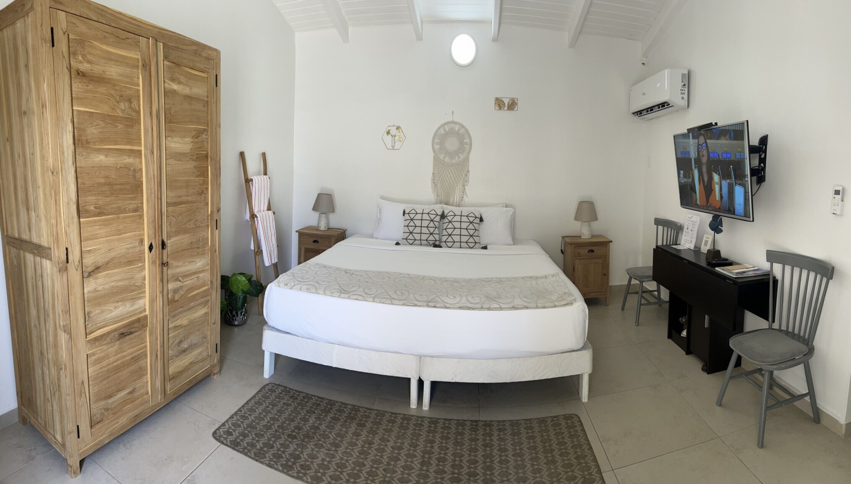 A bedroom with a large wooden wardrobe, a bed with patterned bedding, a desk with a chair, and a TV mounted on the wall.