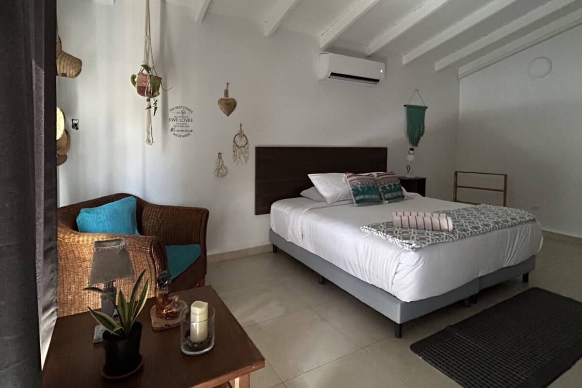 Un dormitorio con cama, mesita de noche, una silla, una pequeña mesa con una planta y un aire acondicionado instalado en la pared.