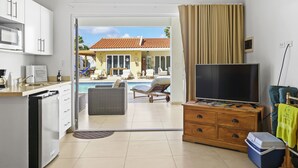 Habitación de hotel con kitchenette, televisión, aparador de madera, maleta y vistas a una piscina y zona de estar al aire libre.