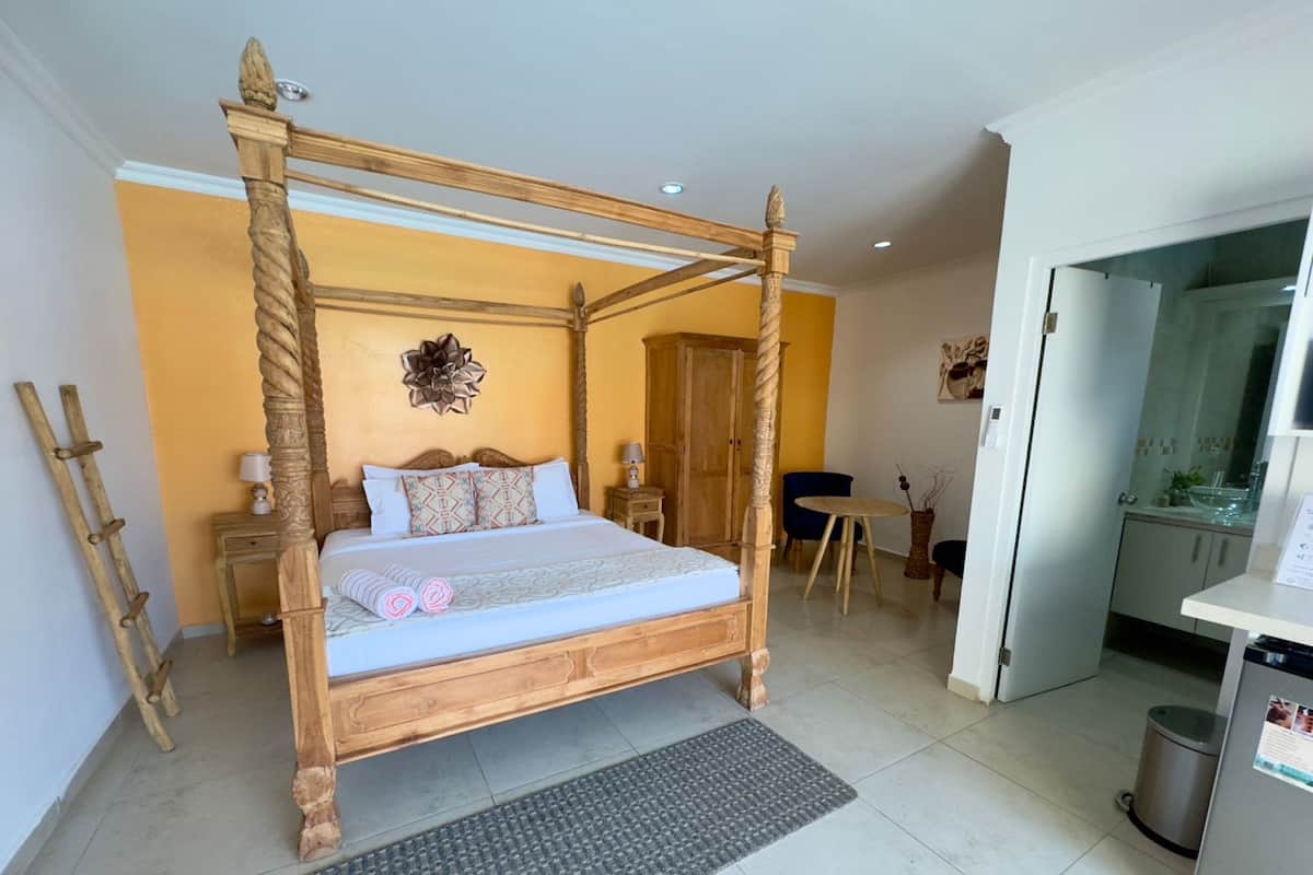 Una cama con dosel de madera, una mesita de noche con lámpara, una silla y una pequeña mesa con un florero.