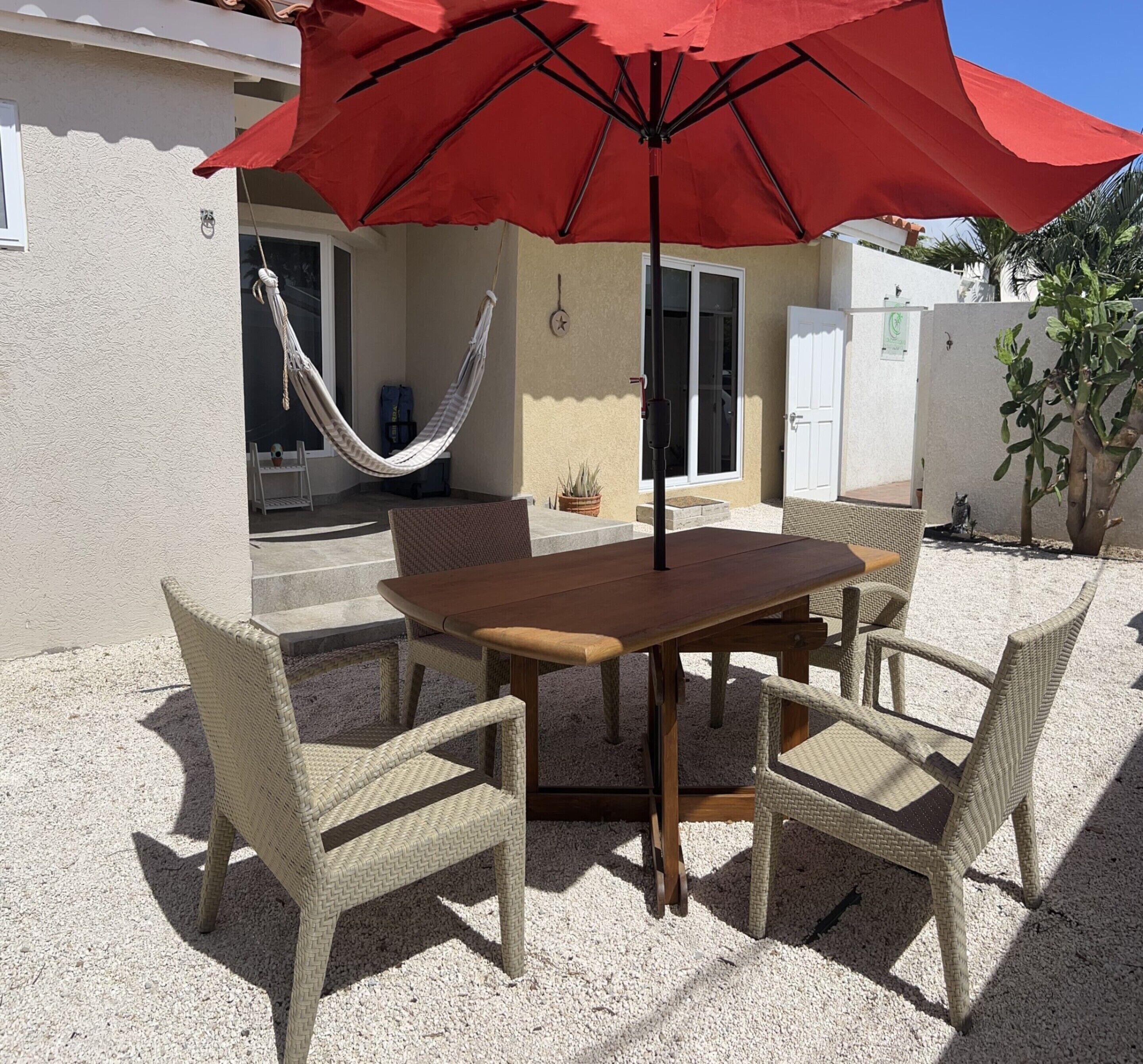 A patio with a wooden table and wicker chairs, a red umbrella, a hammock, and a cactus plant.