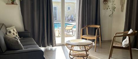 Un salon moderne avec un canapé, une table ronde en osier et deux fauteuils. On y trouve des rideaux, une plante suspendue et une vue sur une piscine à l’extérieur.