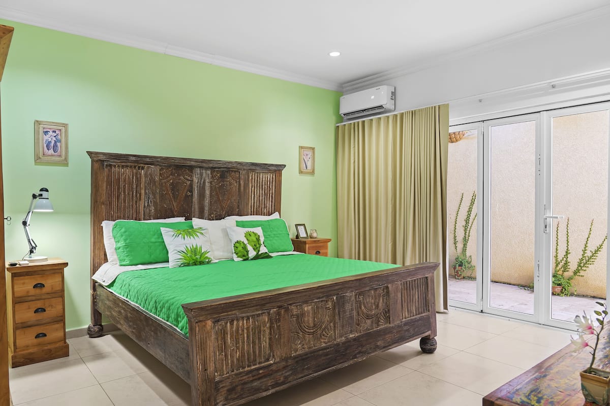 Un dormitorio con cama de madera, ropa de cama verde, una mesita de noche, una lámpara y vistas al exterior a través de una puerta corrediza de vidrio.