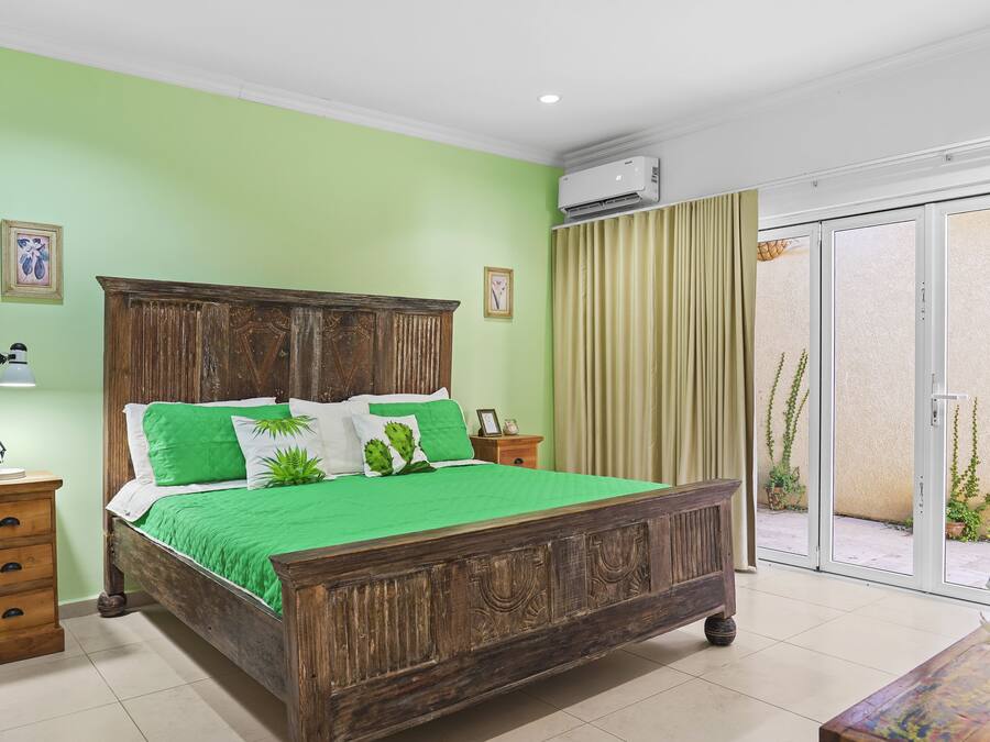 Un dormitorio con cama de madera, ropa de cama verde, una mesita de noche, una lámpara y vistas al exterior a través de una puerta corrediza de vidrio.