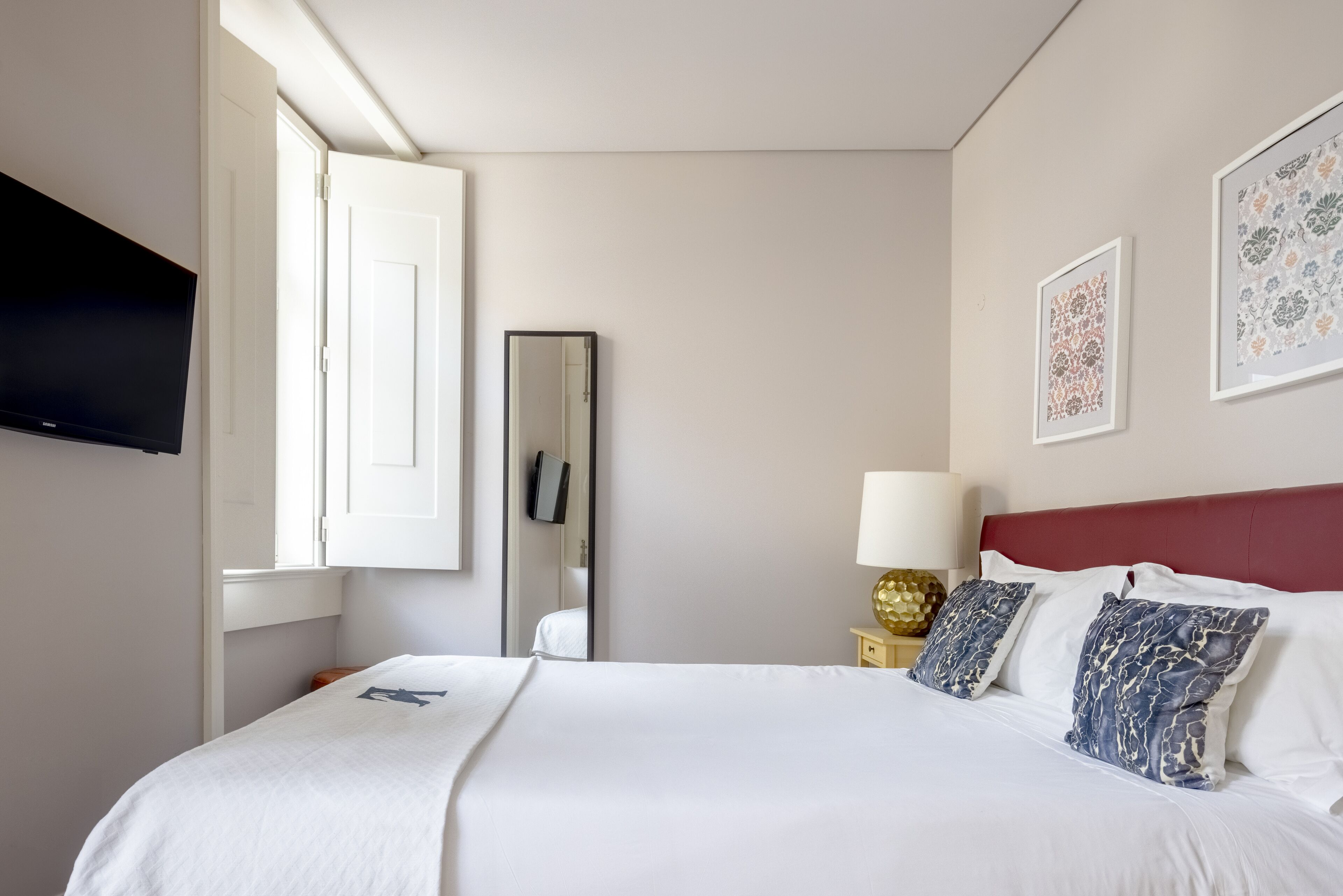 A hotel room with a bed, a TV, a mirror, a bedside table with a lamp, and framed artwork on the wall.