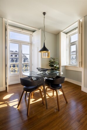 A dining area with a glass table and chairs, a hanging pendant light, and large windows with views of buildings.