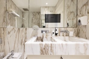A modern bathroom with a marble sink, a handheld mirror, and a wall-mounted soap dispenser.
