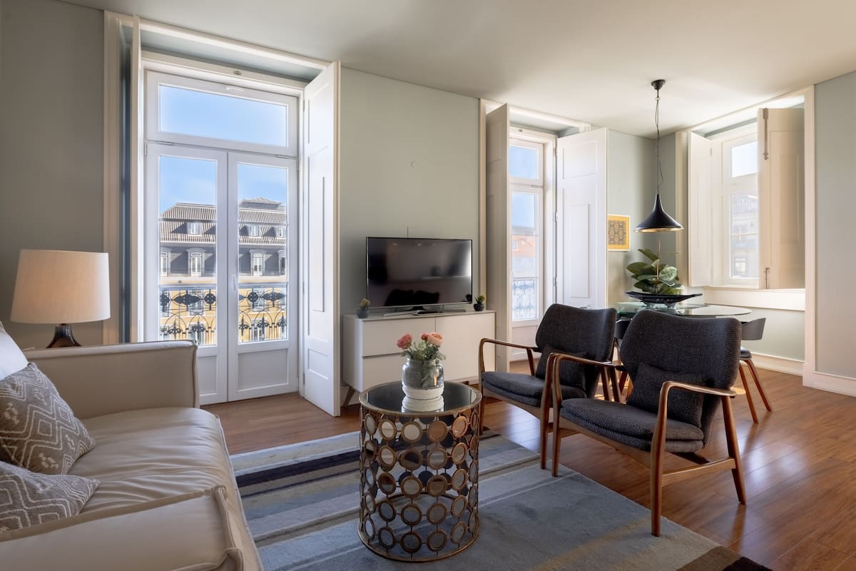 A living room with a sofa, a round coffee table with a decorative base, a flat-screen TV, two armchairs, a dining table with chairs, and large windows with a view of buildings.