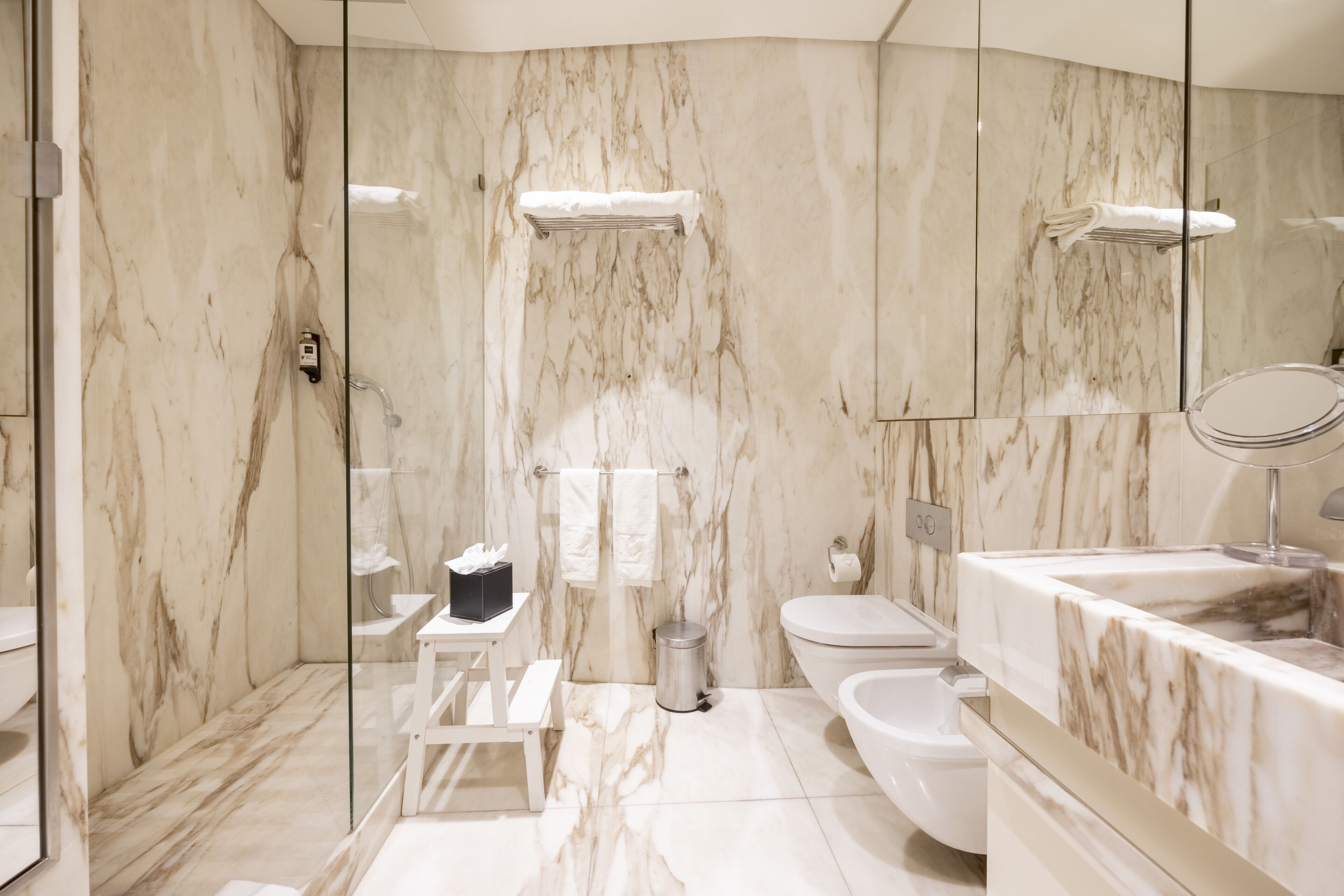 Un bagno moderno con una parete in marmo, box doccia in vetro, un WC, un bidet e un mobile lavabo con specchio.