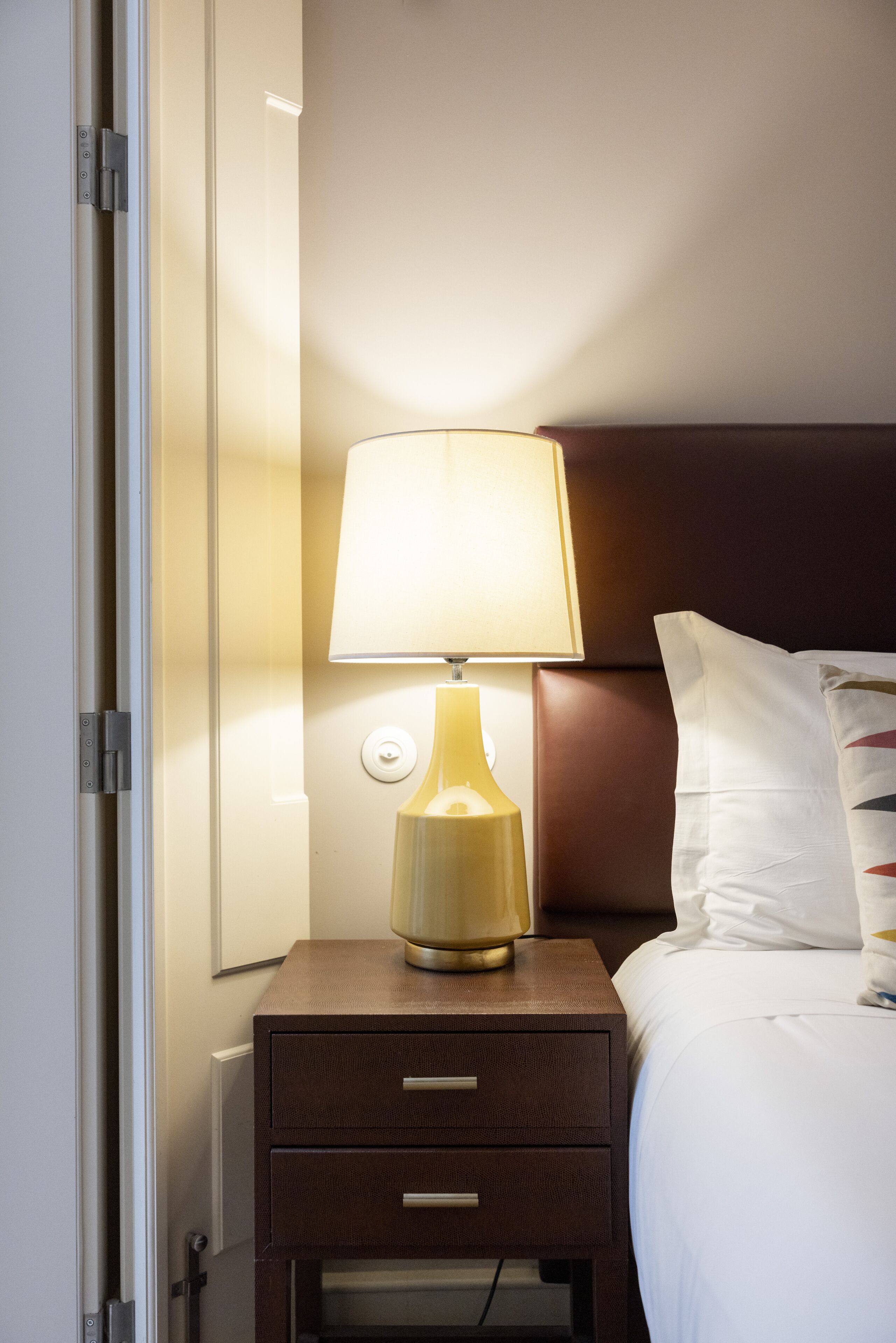 A neatly made bed with white linens, a bedside lamp, and a wooden nightstand with drawers.