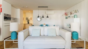 A modern living room with a white sofa, teal accent pillows, and a glass coffee table. There is a kitchen area with a stove and oven visible. - Chiado Square - Lisbon Best Apartments (Lisbon)