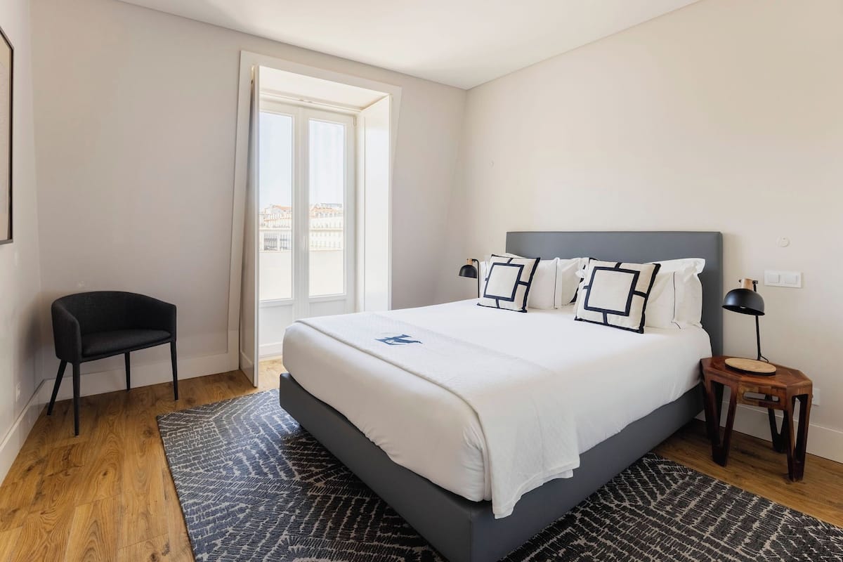 Une chambre à coucher comprenant un lit, une chaise, une table de chevet, une lampe et une vue sur des immeubles par la fenêtre.