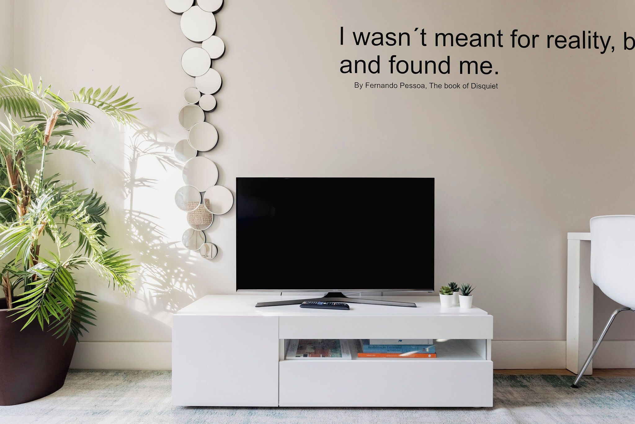 Un salon moderne avec un téléviseur à écran plat posé sur un meuble blanc, une plante en pot et une citation affichée sur le mur.