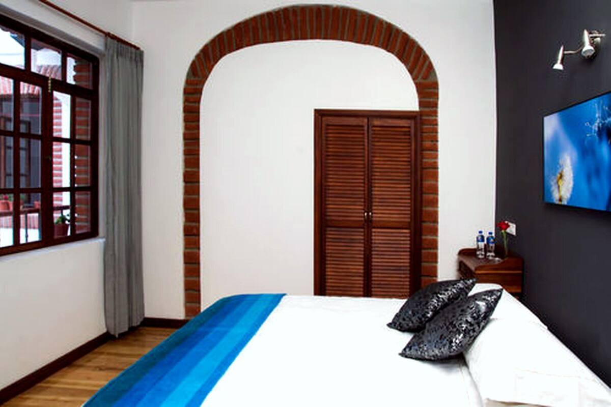 Ein Hotelzimmer mit einem großen Bett, einer Holztür, einem Fenster mit Vorhängen und einem kleinen Tisch mit einer Vase und Flaschen.