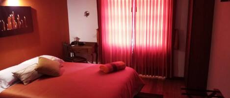A hotel room with a bed, a desk, and a window with red curtains.