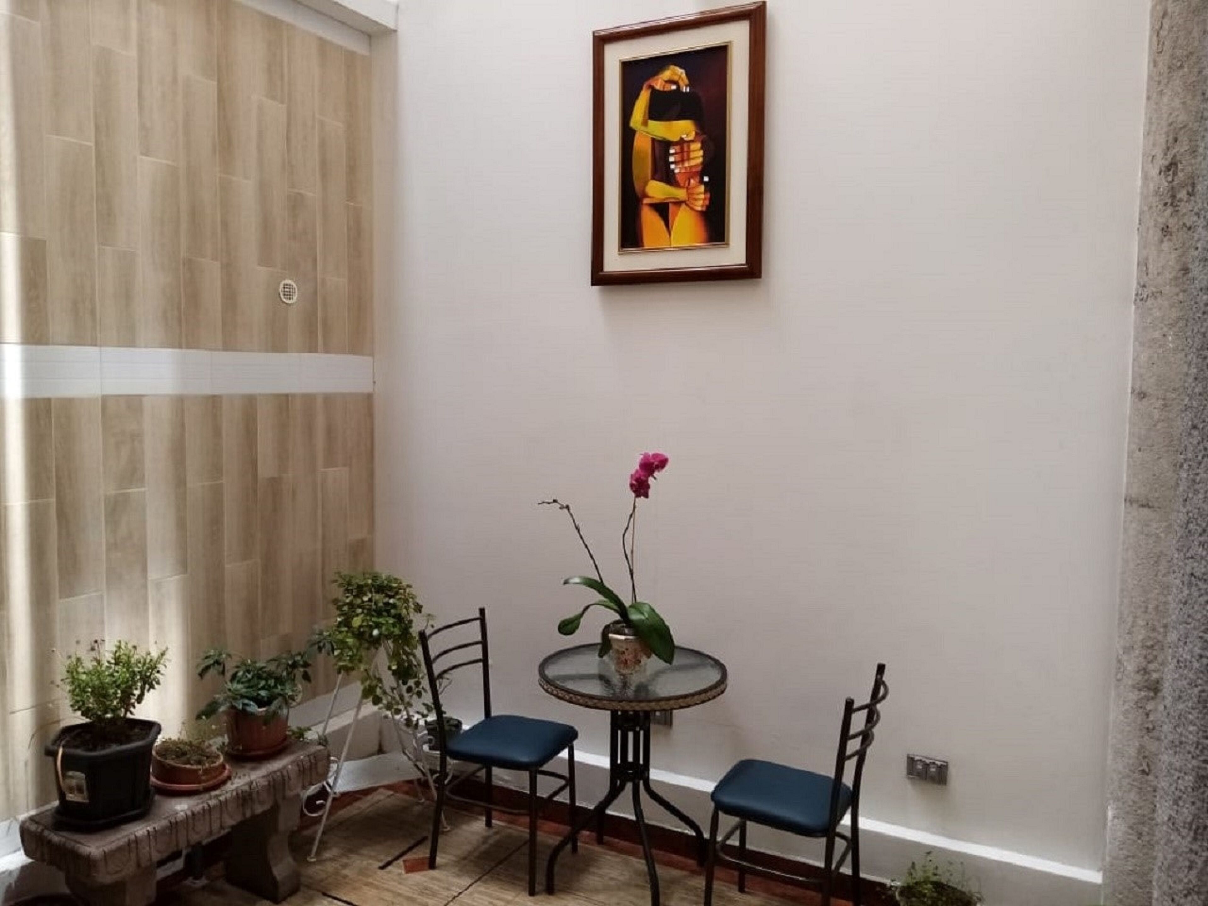 A small, well-lit room with a wooden floor, a round glass table with two chairs, and several potted plants.