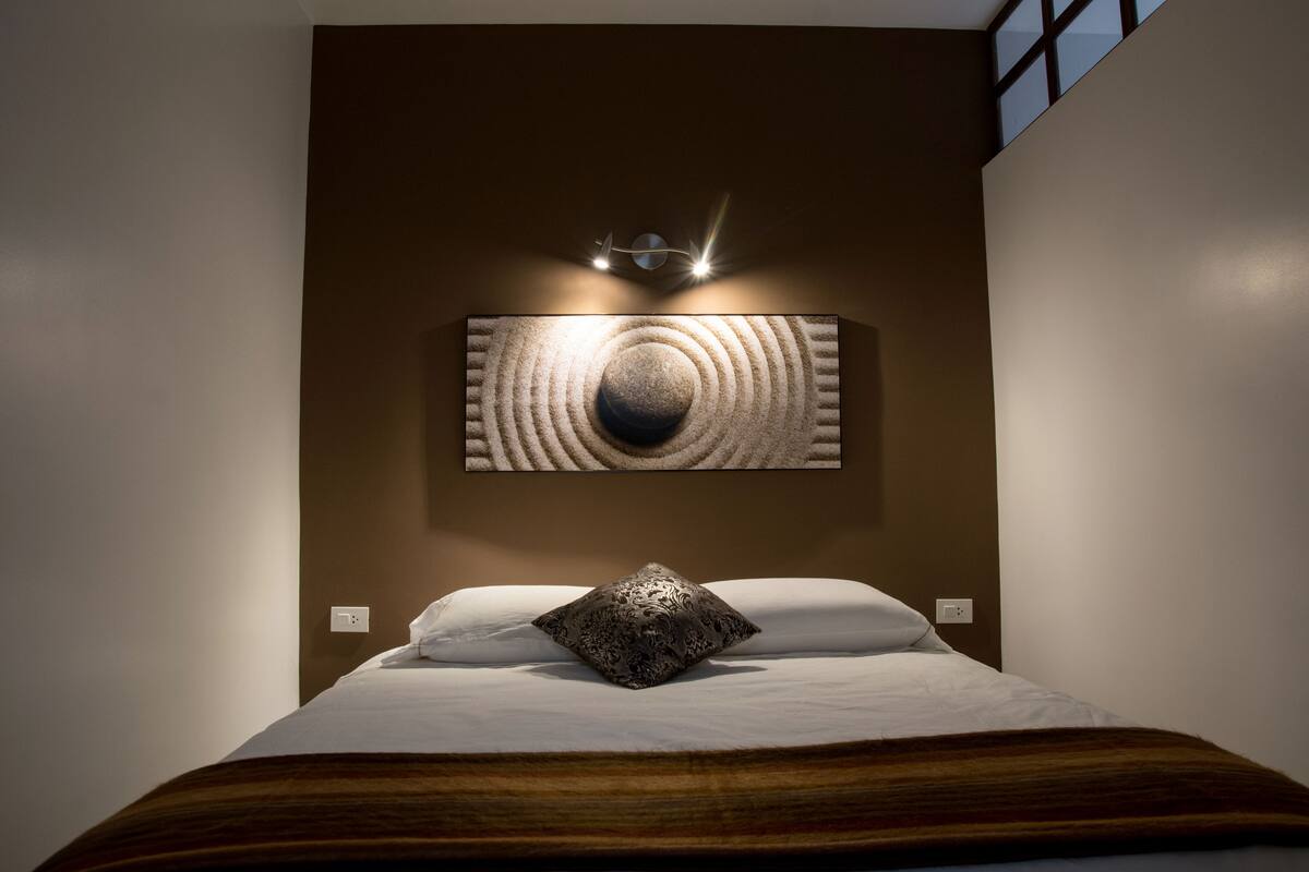 Ein modernes Hotelzimmer mit einem großen Bett, einer dekorativen Wandgestaltung darüber und einem Fenster auf der rechten Seite.
